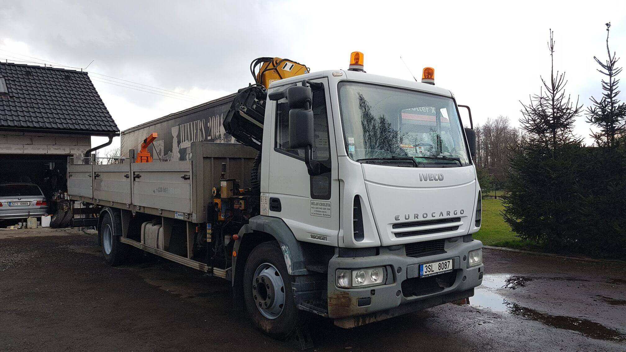 Bílý valník Iveco EuroCargo s hydraulickou rukou zaparkovaný před garáží