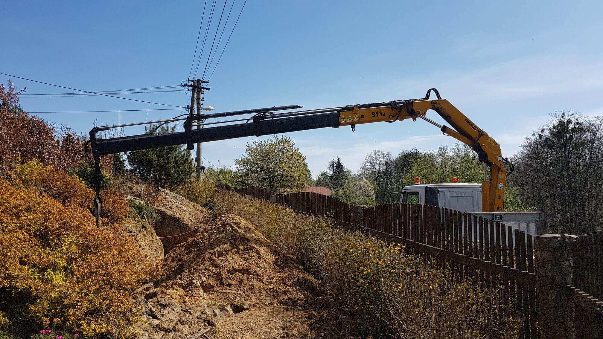 Hydraulická ruka natažená přes dřevěný plot skládá materiál do zahrady
